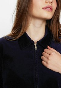 Jūrų laivyno kanalų džemperis su apykakle, priekiniais užtrauktukais ir tekstūruota medžiaga. Modelio ranka laiko apykaklę, atskleidžiant artimą detalę.