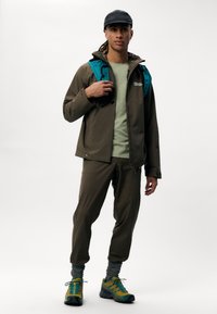Brun vandtæt jakke med teal accenter, grøn t-shirt, matchende brune bukser og multicolor vandresko. Fuldt udendørs outfit.