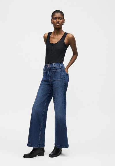 Γυναίκα που φοράει μαύρο αμάνικο τοπ, μπλε τζιν παντελόνι με φαρδιά μπατζάκια και ψηλόμεσο, και μαύρα μποτάκια αστραγάλου, στέκεται με τα χέρια στις τσέπες.