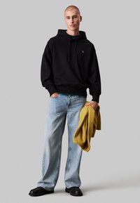 Felpa nera con cappuccio e stringhe, jeans a gamba larga blu chiaro, e un maglione giallo senape appoggiato sul braccio. Scarpe nere completano il look.
