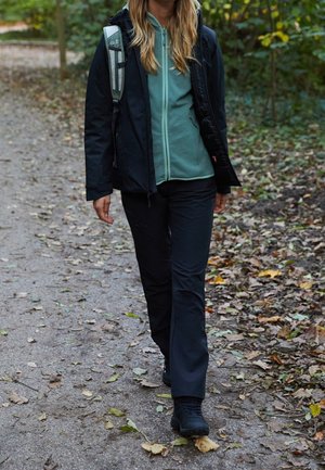 Frau geht auf einem mit Blättern bedeckten Waldweg, trägt schwarze Hose, schwarze Jacke, grünen Kapuzenpullover mit Reißverschluss, schwarze Wanderschuhe und trägt einen Rucksack.