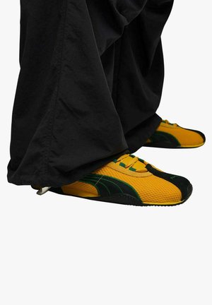 Sneakers Puma in mesh giallo e nero, con dettagli verdi e suola in gomma testurizzata. Pantaloni in tessuto nero coprono parzialmente le scarpe.