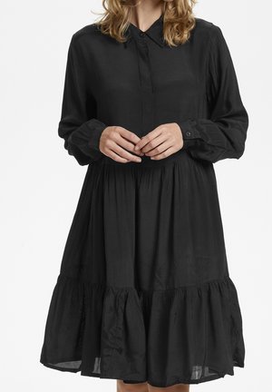 Robe noire à boutonnière fabriquée dans un tissu léger, avec des manches longues, un ourlet volanté et une coupe décontractée. Détail de col simple visible.