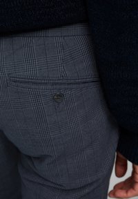 Pantaloni grigi a quadri con una tasca sul retro e dettaglio di bottone. Il tessuto ha una finitura testurizzata, caratterizzata da linee sottili e un design strutturato.