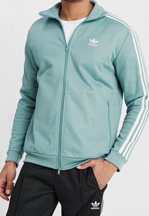 Homme portant une veste zippée Adidas bleu clair avec des bandes blanches sur les manches et un pantalon de survêtement Adidas noir avec logo blanc.