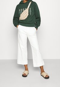 Felpa verde con grafica "SPORTY", pantaloni bianchi a gamba larga, borsa a tracolla beige e sandali beige con cinturini spessi. Tessuti lisci, vestibilità casual.