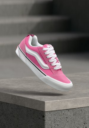 Rózsaszín-fehér alacsony szárú sportcipő fehér fűzőkkel, amely egy betonperem felett lebeg, hátterében szürke lépcsőkkel.