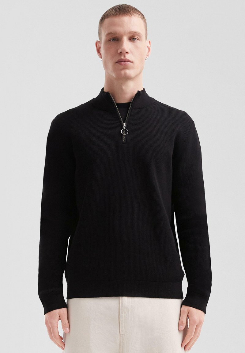 Pull en tricot noir avec un col haut et une fermeture éclair à mi-hauteur. Présente une texture côtelée et des poignets ajustés.