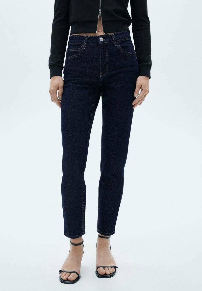 Personne portant un jean skinny taille haute bleu foncé, un top zippé court noir et des talons ouverts à brides noirs sur fond blanc uni.