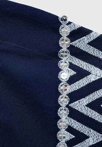 Mornarska tkanina s bijelim izvezenim cik-cak uzorcima i metalnim kružnim detaljima uz jedan rub, dodajući teksturu i detalje.