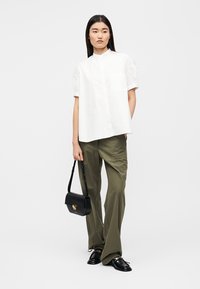 Camicia bianca a maniche corte con colletto e taschino, abbinata a pantaloni a gamba larga color verde oliva. La modella tiene una borsa nera con dettagli dorati.