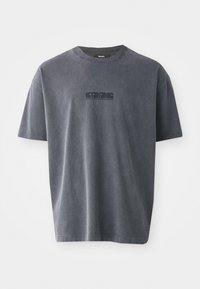 Camiseta gris oscuro de algodón, con cuello redondo, mangas cortas y un logo gráfico negro en el frente. Textura suave y ligeramente desgastada.
