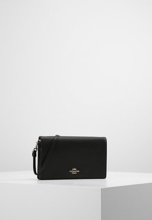 Sort læder Coach håndledspung med gyldigt logo og aftagelig rem, placeret på en hvid piedestal mod en ensfarvet grå baggrund.