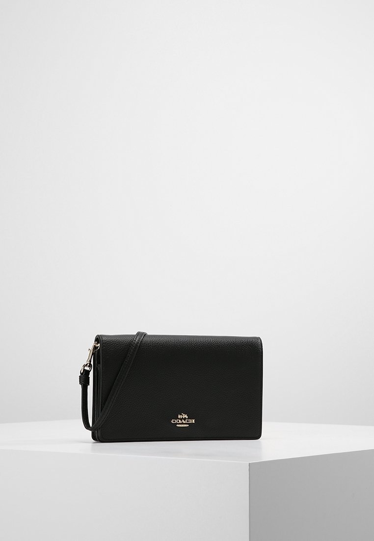 Pochette Coach en cuir noir avec logo doré et bracelet amovible, posée sur un socle blanc sur un fond gris uni.