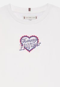 Άσπρη μπλούζα T-shirt με κεντημένο λογότυπο καρδιάς σε κόκκινο, άσπρο και μπλε χρώμα, με το κείμενο "Tommy Hilfiger" κεντραρισμένο κάτω από τον λαιμό.