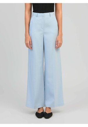 Femme portant un pantalon large taille haute bleu clair associé à un haut noir sans manches et des chaussures pointues noires, debout immobile.