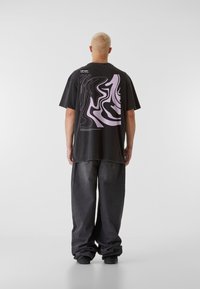 Černé oversized tričko s vlnitým levandulovým grafickým designem na zadní straně, doplněné o široké tmavě šedé džíny.