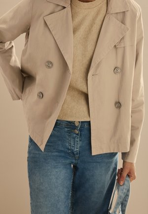 Personne portant une veste beige croisée sur un pull marron clair et un jean bleu, tenant un masque facial en denim dans la main droite.