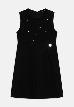 Vestido negro sin mangas con pequeños adornos similares a perlas en el pecho y un pequeño detalle luminoso en forma de corazón cerca de la cintura.