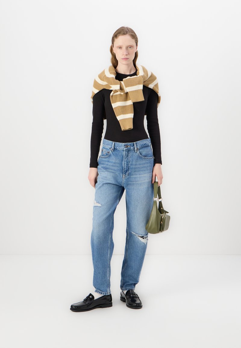 Haut à manches longues noir, pull en tricot rayé beige et blanc noué au cou, jeans bleus clairs déchirés, mocassins noirs et sac à main vert.