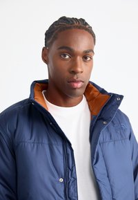 Puffer jacket, blue outer material, orange inner lining, zip closure, high collar, logo detail on left chest, smooth texture.