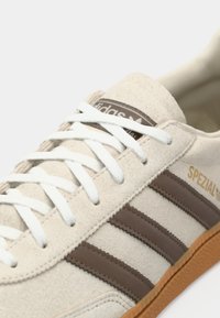 Sneaker in suede beige chiaro con tre strisce marroni e una suola in gomma testurizzata. Presenta lacci bianchi e un'etichetta con logo marrone sulla linguetta.