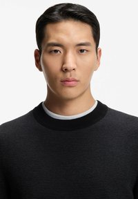 Schwarzer Baumwollpullover mit strukturierter Webart und geripptem Rundhalsausschnitt, über einem weißen Hemd getragen. Glatte Oberfläche, minimale Akzente.