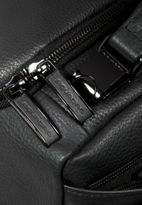 Zwarte leren tas met zichtbare ritsen en elegante metalen details. De tas heeft een gestructureerd oppervlak en subtiele merkaccenten.