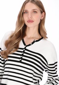 Pull rayé noir et blanc avec une texture côtelée, présentant un col en V et une patte de boutonnage ornée d'accents en perle.