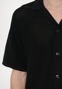 Juodas megztas trumpomis rankovėmis marškinėliai su mygtukų užsegimu, apykakle ir tekstūruota struktūra su atvirų audinio detalių elementais.