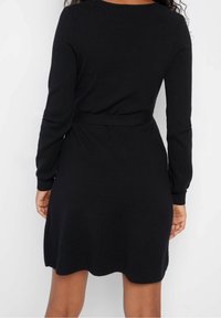 Robe en maille noire à manches longues, col arrondi, taille ceinturée et ourlet évasé. Texture douce et finition lisse sans motifs visibles.