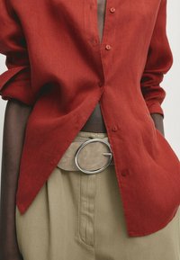 Chemise rouge en lin à boutons avec manches retroussées, associée à un pantalon beige et une large ceinture avec une boucle circulaire argentée.