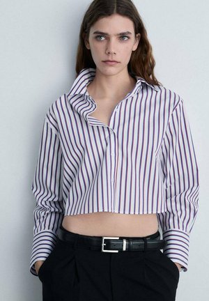 Camicia bianca tagliata con righe verticali viola, colletto ampio e chiusura a bottoni, abbinata a pantaloni neri a vita alta e una cintura nera.
