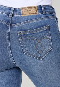 Jean en denim bleu à taille haute, avec une étiquette beige à l'arrière, des coutures élaborées sur les poches et une coupe classique.