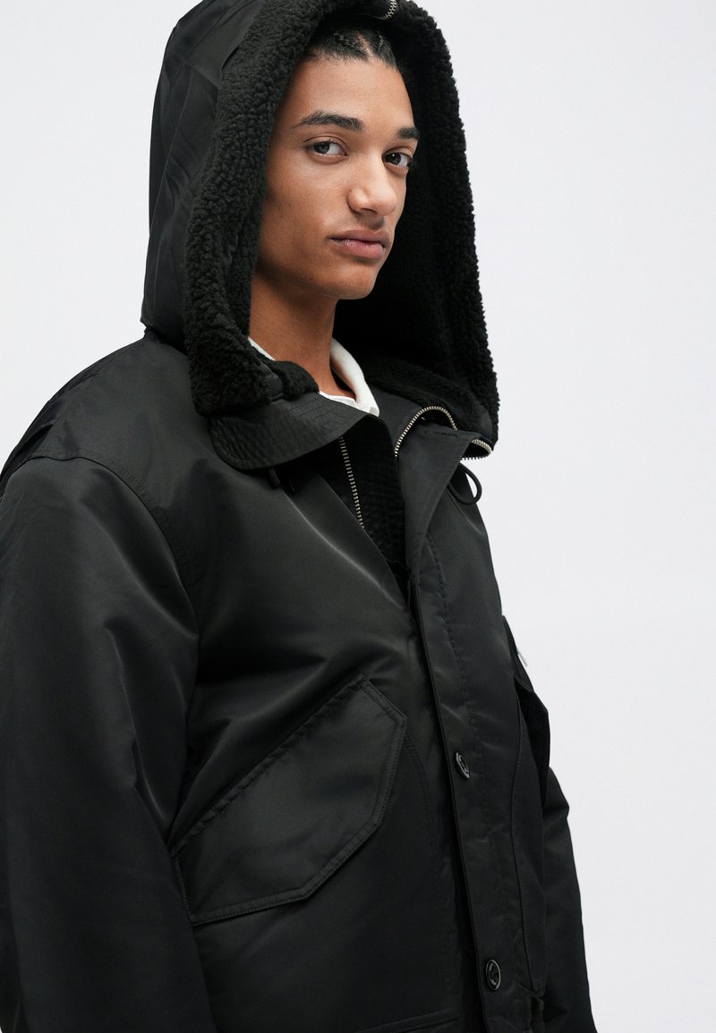 Veste extérieure noire avec une doublure en peluche pour la capuche, dotée de grandes poches avant et d'une fermeture éclair. Le tissu semble lisse et résistant à l'eau.
