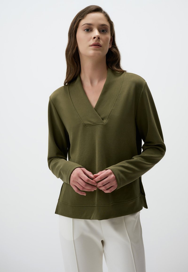 Maglia verde oliva a maniche lunghe con scollatura a V, texture morbida e spacchi laterali, abbinata a pantaloni color crema.
