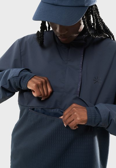 Marinblå hoodie-jacka med en dragkedjelombå, texturerat tyg och ståkrage. Har långa ärmar och en matchande keps.