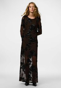 Robe maxi en dentelle noire à fleurs avec manches longues, ornée d'appliqués floraux 3D texturés sur une doublure transparente ajustée.