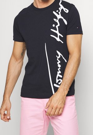 Mężczyzna ubrany w czarną koszulkę Tommy Hilfiger z dużym białym logo oraz jasnoróżowe szorty, stojący na jednolitym tle.