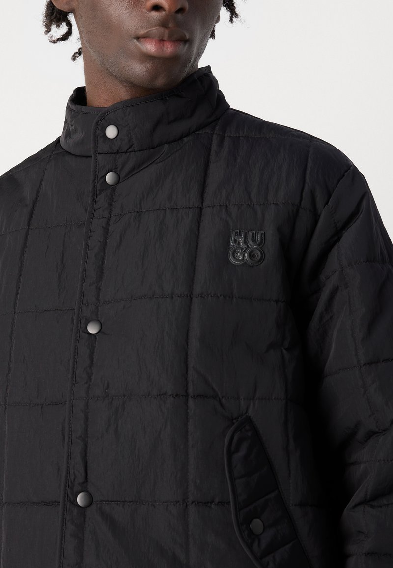 Veste matelassée noire en tissu léger, avec un col haut, des boutons-pression et un patch logo sur la poitrine gauche.