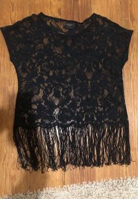 Svart spetsblus med korta ärmar, prydd med blommönster och fransar i nederkant. Tyget har en genomskinlig struktur och en mjuk hängande form.