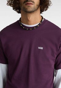 Fialové bavlněné tričko s bílým logem "VANS" na hrudi, vrstvené přes bílé tričko s dlouhým rukávem, s korálkovým náhrdelníkem.