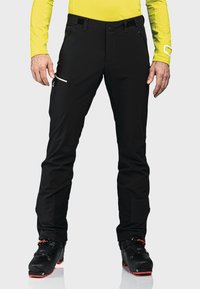 Schwarze Skihosen mit schlanker Passform, ausgestattet mit seitlichen Reißverschlusstaschen und einer glatten Textur. Für optimale Leistung entworfen, kombiniert mit schwarzen Skischuhen.