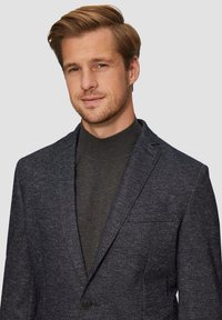 Navyfarbener strukturierter Blazer mit Reverskragen, Einknopfverschluss und Seitentasche, getragen über einem dunkelgrauen Rollkragenpullover.