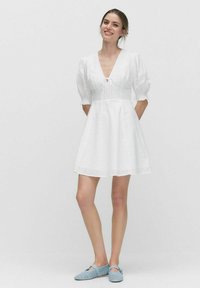 Robe blanche courte avec des manches bouffantes, un col en V avec un lien, et une jupe fluide. Le tissu a une texture subtile. Portée avec des chaussures bleu clair.