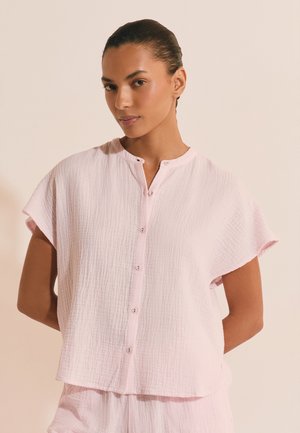 Kobieta z zaczesanymi do tyłu włosami, ubrana w jasno-różowy, teksturowany, krótki rękaw z guzikami i pasujące szorty, stojąca na beżowym tle.