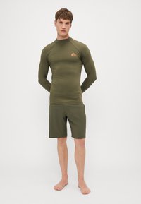 Alyvuogių spalvos rash guard su ilgomis rankovėmis ir aukštu apykakle, derinamas su atitinkančiomis šortais. Apima prekės ženklo logotipą ant krūtinės ir kairės rankovės.