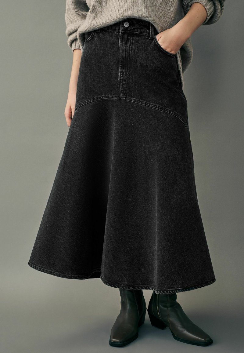 Gonna midi in denim nero con design svasato, dotata di tasche laterali e una vita visibile. Abbinata a stivali di caviglia scuri.