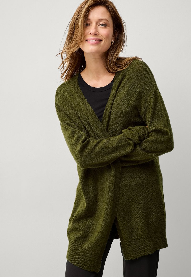 Cardigan verde a maglia con vestibilità ampia, scollatura a V, maniche lunghe e spacchi laterali, indossato sopra una maglietta nera. Texture morbida, design casual.