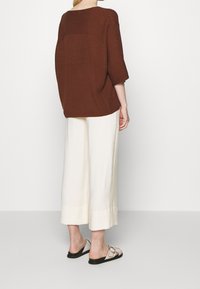 Maglione oversize marrone lavorato a maglia, pantaloni a gamba larga crema e sandali neri. Il maglione presenta un dettaglio di cucitura testurizzata sulla schiena.